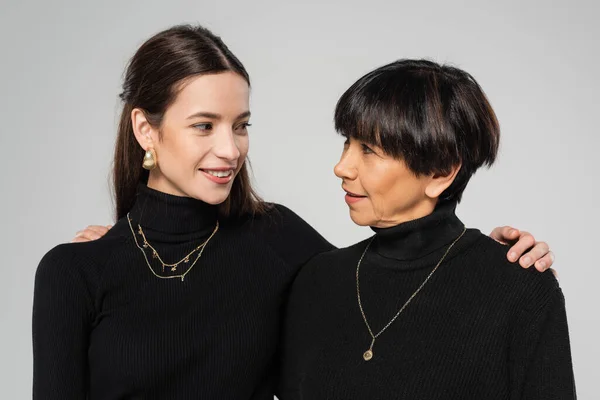 Brunette asian mother and daughter in black turtlenecks and necklaces smiling at each other isolated on grey — Fotografia de Stock