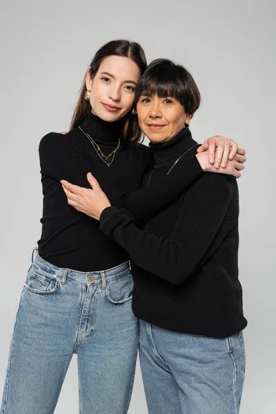 Мать и дочь азиатки в черных водолазках и джинсах обнимаются и смотрят на камеру, изолированную на сером — стоковое фото