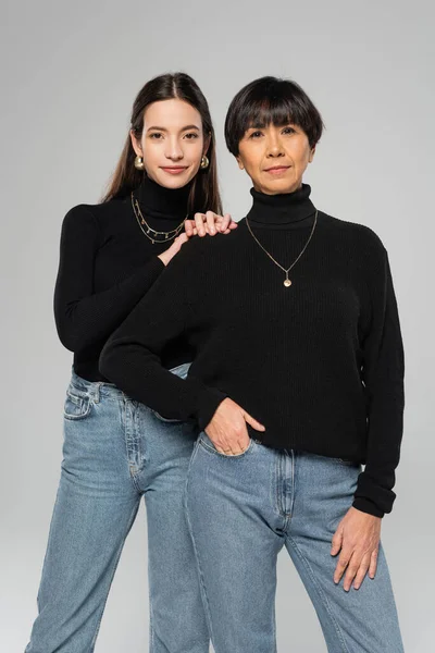 Стильная азиатская мать и дочь в черных водолазках и синих джинсах позируют изолированно на сером — стоковое фото