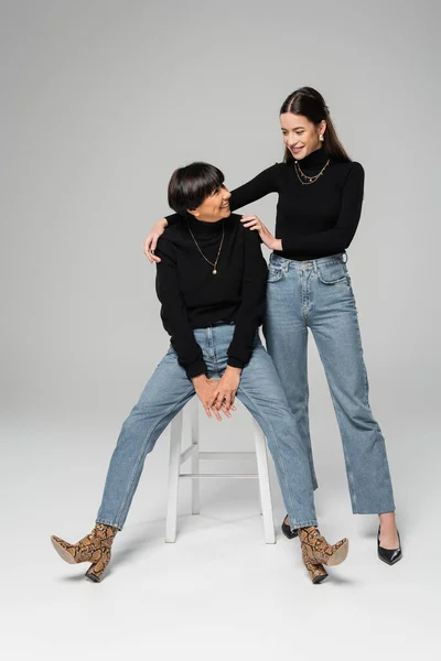 Full length of stylish woman hugging shoulders of cheerful asian mother sitting on chair on grey background — Stock Photo