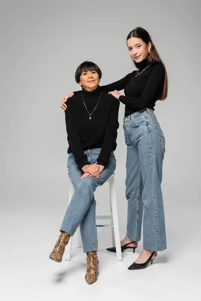 Full length of happy asian woman hugging shoulders of middle aged mother sitting on chair on grey background — Stock Photo