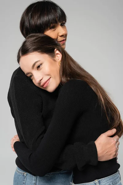 Pleased asian mother and daughter in black turtlenecks embracing isolated on grey — Stockfoto