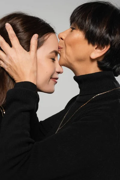 Vue latérale d'une femme asiatique d'âge moyen au col roulé noir embrassant le front d'une jeune fille isolée sur fond gris — Photo de stock