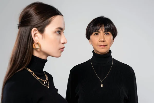Brunette asian woman in black turtleneck looking at camera near blurred daughter in jewelry isolated on grey — Fotografia de Stock