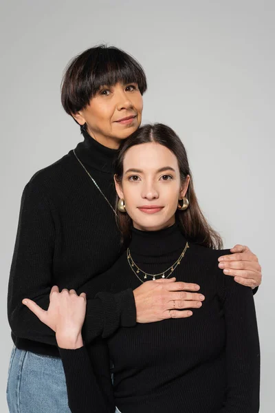 Smiling asian woman looking at camera and hugging stylish daughter wearing black turtleneck isolated on grey — Stock Photo