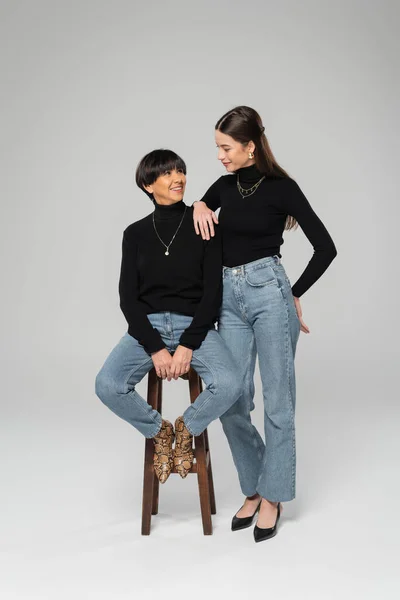Young woman in black turtleneck and jeans smiling at trendy asian mother sitting on wooden stool on grey background — Stock Photo