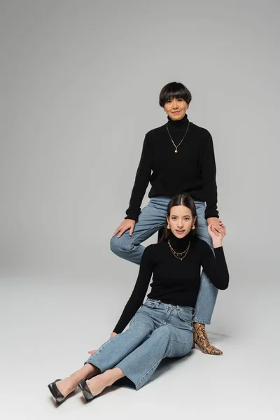Full length of trendy asian mother and daughter in jeans and black turtlenecks holding hands while posing on grey background — Stockfoto