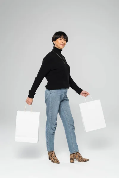 Longitud completa de morena mujer asiática en cuello alto negro y vaqueros azules posando con bolsas de compras blancas sobre fondo gris - foto de stock