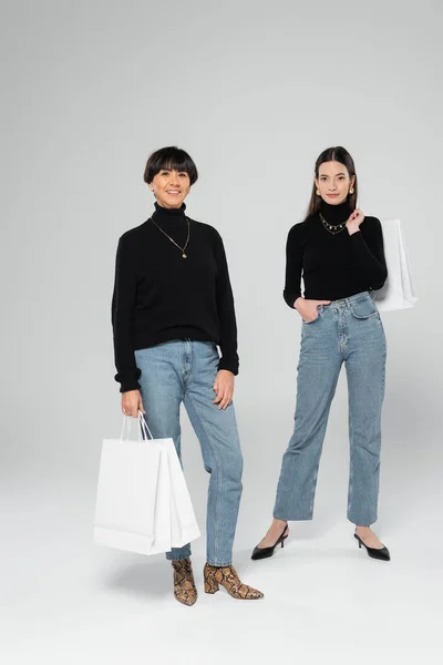 Полная длина азиатской матери и дочери в модном наряде позируя с белыми сумками покупок на сером фоне — стоковое фото