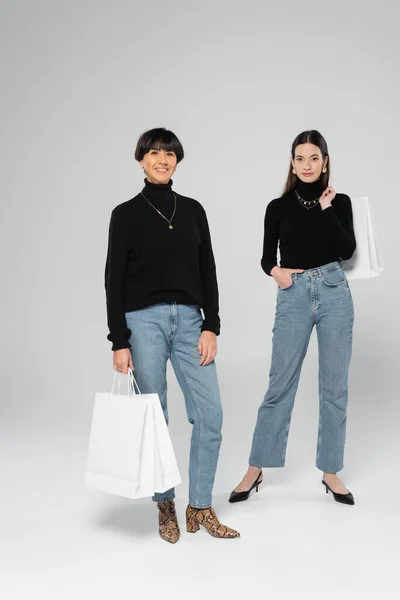 Piena lunghezza di sorridente asiatica madre e figlia in jeans e dolcevita nero in posa con borse della spesa bianche su sfondo grigio — Foto stock