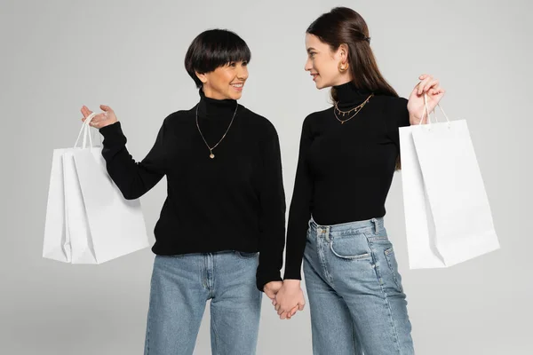 Fashionable asian mother and daughter holding hands and smiling at each other while holding shopping bags isolated on grey - foto de stock