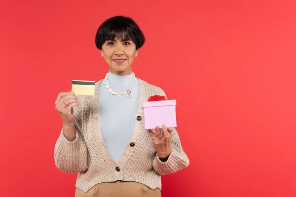 Donna asiatica di mezza età in maglia cardigan contenente scatola regalo e carta di credito isolata su corallo — Foto stock