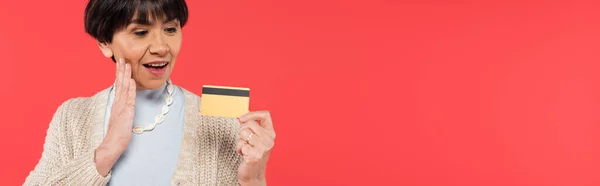 Impressed asian woman in cardigan touching face while holding credit card isolated on coral, banner — Photo de stock