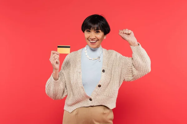 Bruna asiatico donna in maglia cardigan holding carta di credito e celebrare il successo isolato su corallo — Foto stock