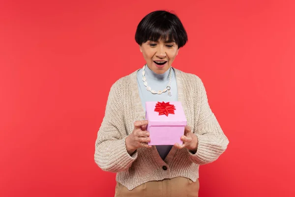 Sorpreso donna asiatica in maglia cardigan guardando scatola regalo con fiocco rosso isolato su corallo — Foto stock