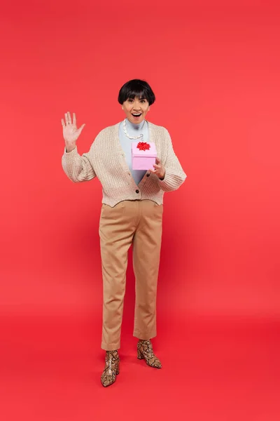 Full length of excited asian woman in stylish clothes holding present and waving hand on coral background — Photo de stock