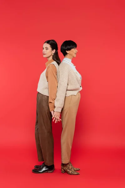 Full length of asian mother and daughter in stylish casual attire holding hands while standing back to back on coral background — Stock Photo