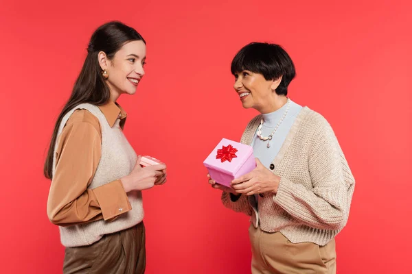 Pleased asian mother and daughter in stylish clothes holding presents and looking at each other isolated on coral — Stock Photo