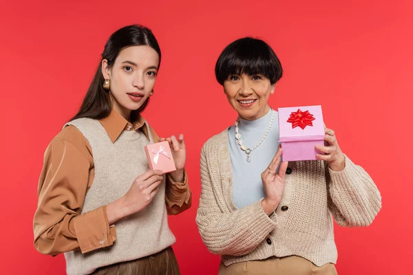 Stylish asian mother and daughter showing presents and smiling at camera isolated on coral — Stock Photo