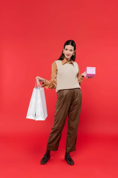 Volle Länge der glücklichen Frau in trendiger Freizeitkleidung posiert mit Einkaufstaschen und Geschenkschachtel auf Korallenhintergrund — Stockfoto