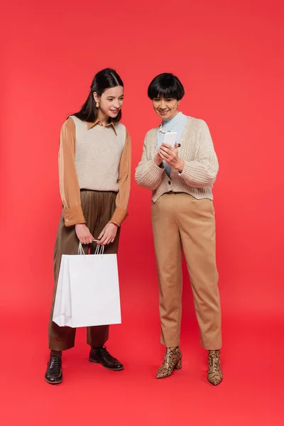 Longitud completa de la elegante mujer asiática mostrando el teléfono celular a la hija sonriente sosteniendo bolsas de compras en el fondo de coral - foto de stock