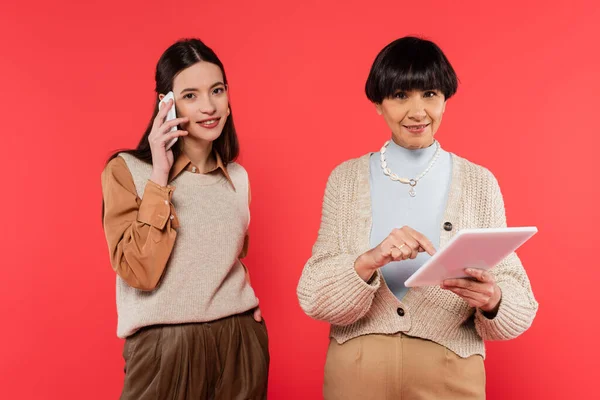 Young asian woman talking on mobile phone near smiling mother using digital tablet isolated on coral — Foto stock