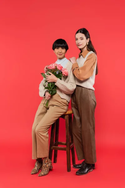 Full length of young asian daughter hugging happy mother sitting on high chair and holding bouquet of flowers on coral — Foto stock