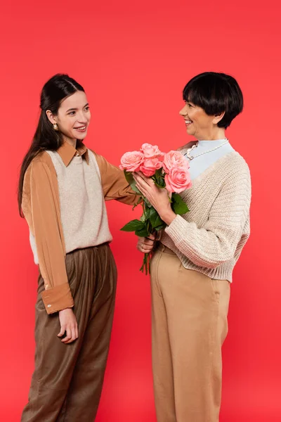 Happy asian daughter looking at cheerful mother with flowers in hands isolated on coral — Stock Photo