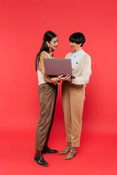 Full length of happy asian mother and daughter holding laptop on coral background — Foto stock