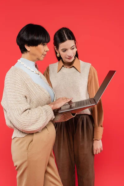 Donna di mezza età utilizzando laptop vicino a giovane figlia asiatica isolata sul corallo — Foto stock