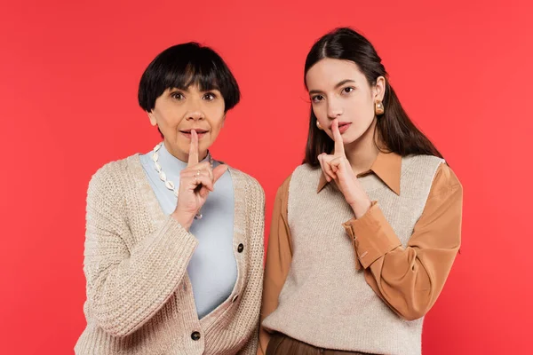 Brunette asian mother and daughter in stylish clothes showing hush sign isolated on coral — Stock Photo