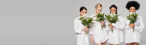 Young multiethnic women wearing white shirts and smelling aromatic flowers isolated on grey, banner — Stock Photo