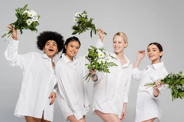 Возбужденные мультикультурные женщины в рубашках, держащие букеты белых цветов с зелеными листьями, изолированными на сером — стоковое фото