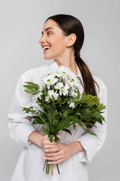 Joyeuse femme brune en chemise blanche tenant des fleurs fraîches et regardant loin isolé sur gris — Photo de stock