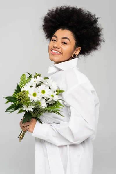 Curly african american woman in white shirt holding spring bouquet and smiling at camera isolated on grey — Stock Photo