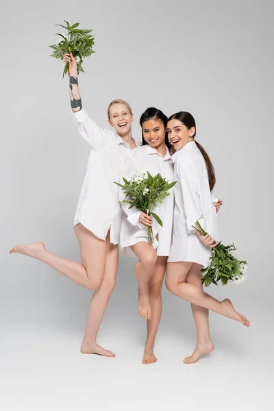Полная длина счастливой татуированной модели держа цветы в поднятой руке рядом с босыми межрасовыми женщинами в белых рубашках на сером фоне — стоковое фото