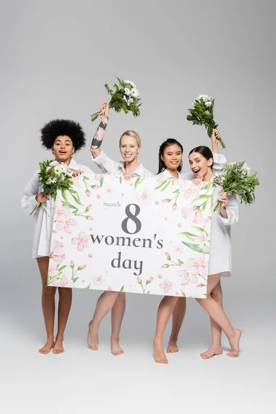 Full length of cheerful multiethnic women holding flowers and greeting placard with womens day lettering on grey background — Stock Photo