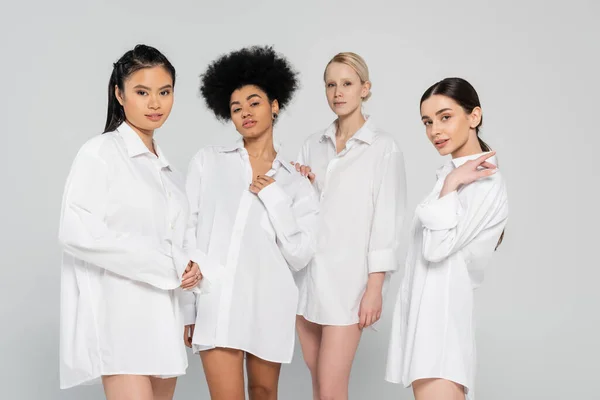 Pretty and sensual multiethnic models in white shirts looking at camera isolated on grey — Photo de stock