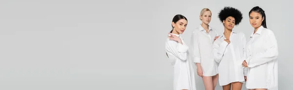 Sensual multiethnic women in white shirts looking at camera isolated on grey, banner — Foto stock