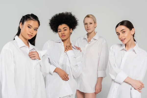 Young multiethnic women looking at camera while posing in white shirts isolated on grey — Foto stock