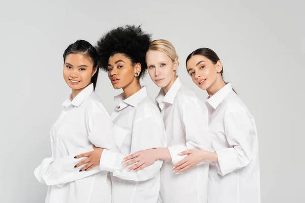 Молоді багатоетнічні жінки дивляться на камеру, позуючи в білих сорочках ізольовані на сірому — стокове фото