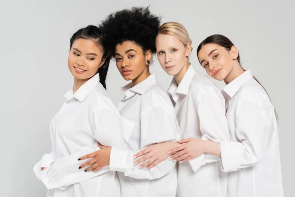Позитивні і чуттєві мультикультурні жінки дивляться на камеру, позуючи в білих сорочках ізольовані на сірому — стокове фото