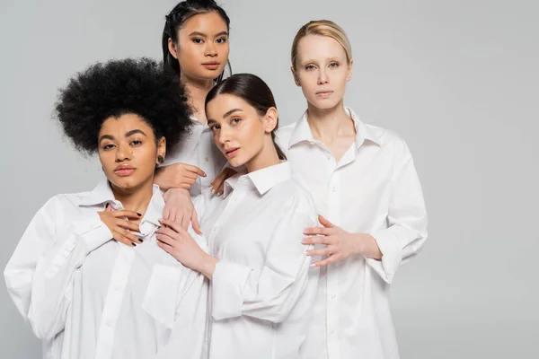 Sensual multicultural women in white shirts looking at camera while standing isolated on grey — Stockfoto