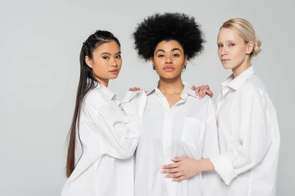 Молоді багатокультурні жінки в білих сорочках торкаються плечей афроамериканського зразка, ізольованого на сірих. — стокове фото
