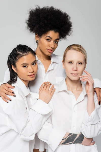 Curly african american woman hugging shoulders of young multiethnic models in white shirts isolated on grey — Stock Photo
