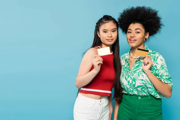 Young and stylish interracial women showing business and credit cards isolated on blue - foto de stock