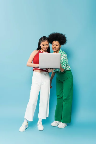 Full length of young multiethnic woman in trendy clothes smiling while looking at laptop on blue background — Foto stock