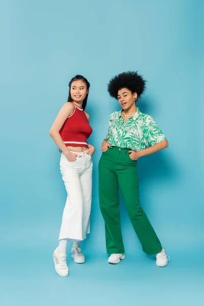 Full length of asian and african american women in fashionable clothes posing with hands in pockets on blue background — Stock Photo