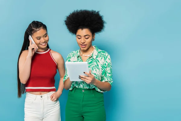 Cheerful asian woman talking on smartphone near african american friend with digital tablet isolated on blue — Stock Photo