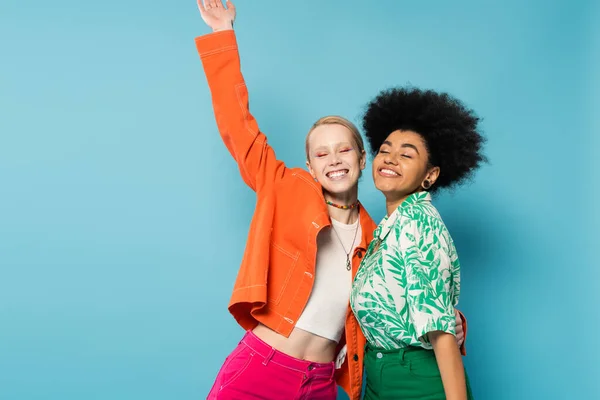 Happy interracial women in colorful clothes posing with closed eyes isolated on blue — Stock Photo